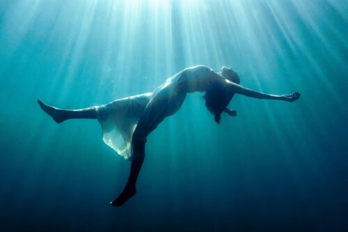 A woman underwater in a position of letting go and floating upwards, with sun rays from above shining through her. A transcendental image.