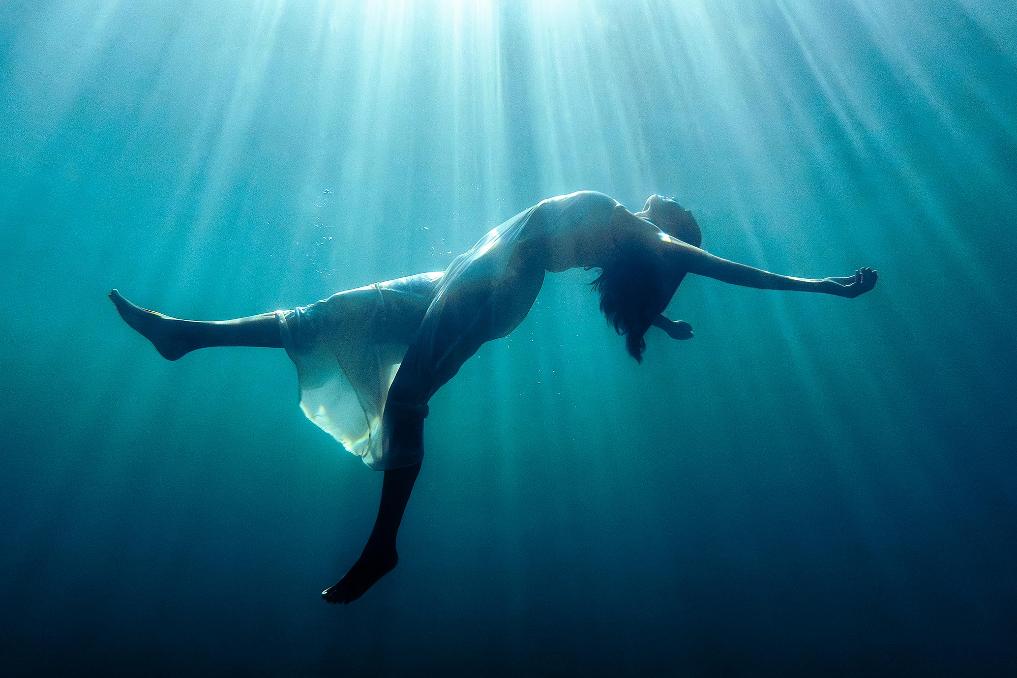 A woman underwater in a position of letting go and floating upwards, with sun rays from above shining through her. A transcendental image.