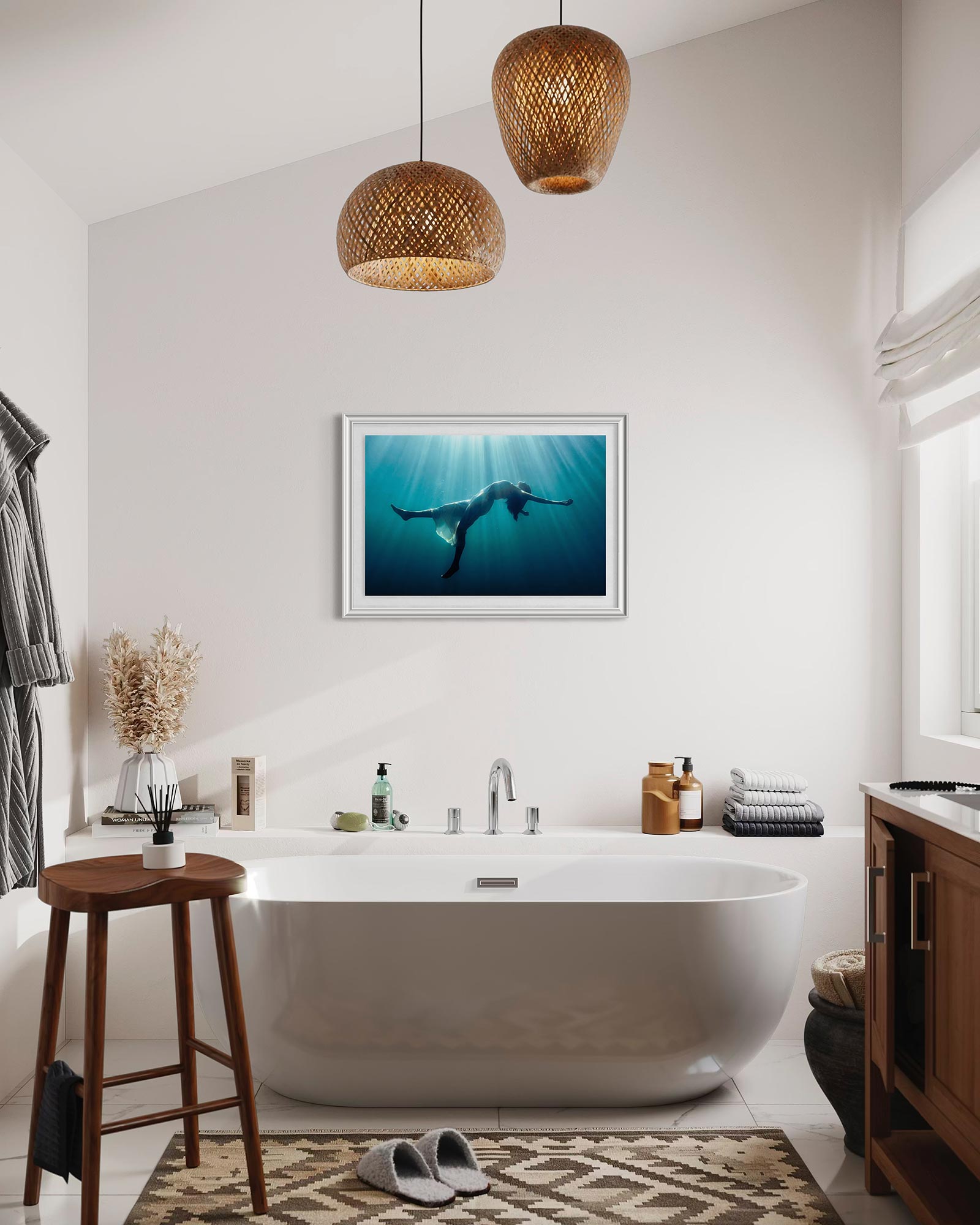 A framed underwater fine art photograph displayed above a freestanding bathtub in a bright, minimalist bathroom, showing a floating female figure suspended in blue water with soft light rays.