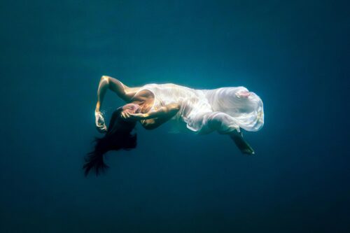 A woman floating underwater in a flowing white dress, suspended in deep blue water, her body gently arched in a moment of stillness and weightlessness.