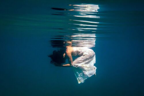 Underwater fine art photograph of a woman curled gently beneath the water surface, wearing a flowing white dress, with soft reflections and deep blue tones.