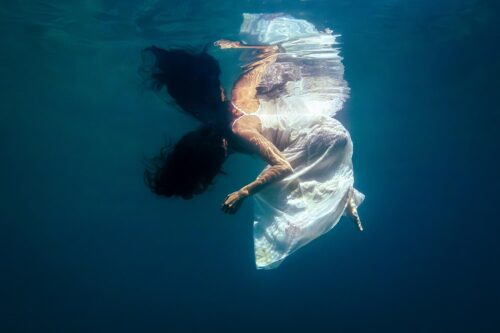 Underwater fine art photograph of a woman rising toward the water surface, wearing a flowing white dress, with soft light and deep blue tones.