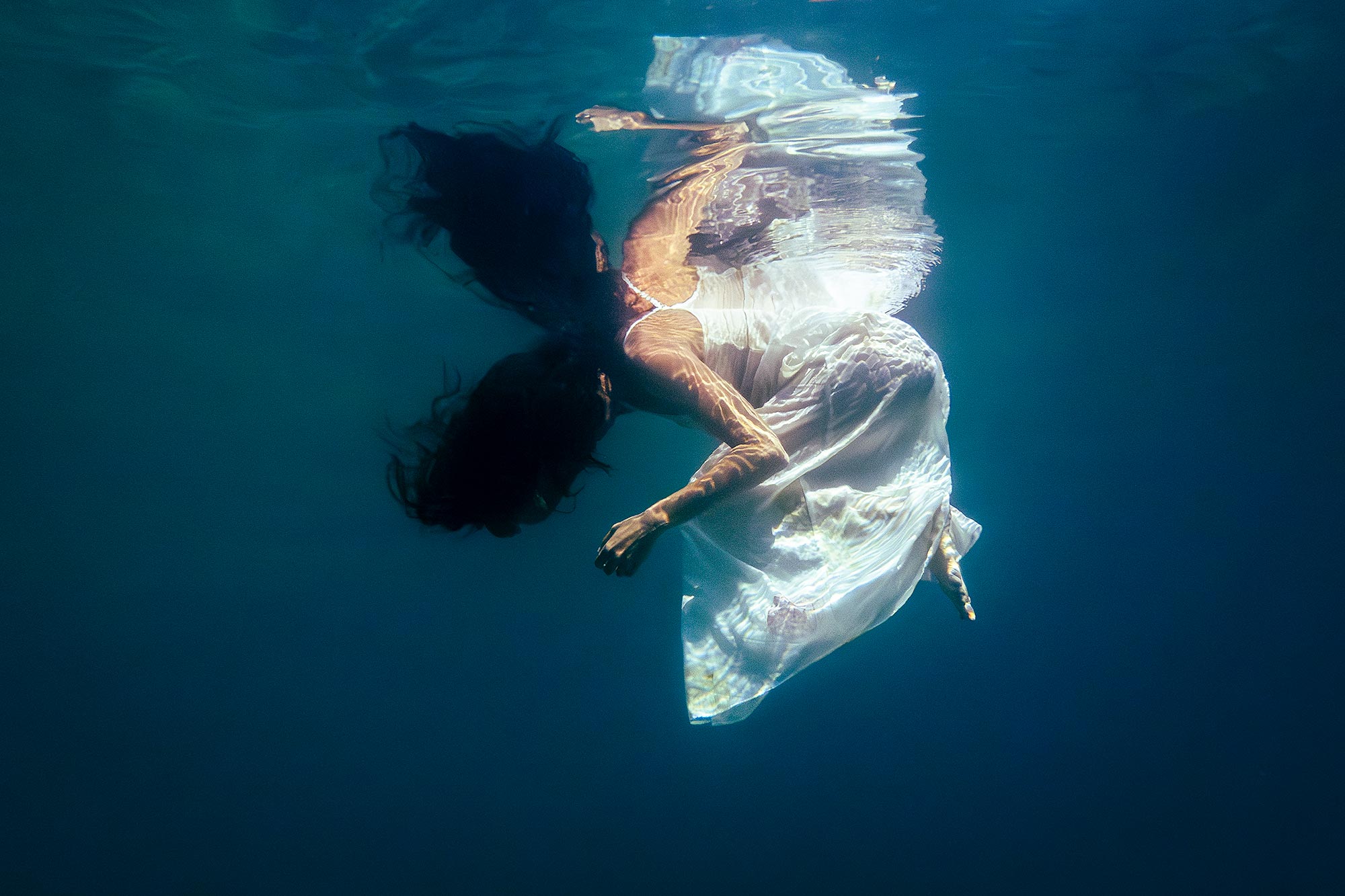 Underwater fine art photograph of a woman rising toward the water surface, wearing a flowing white dress, with soft light and deep blue tones.