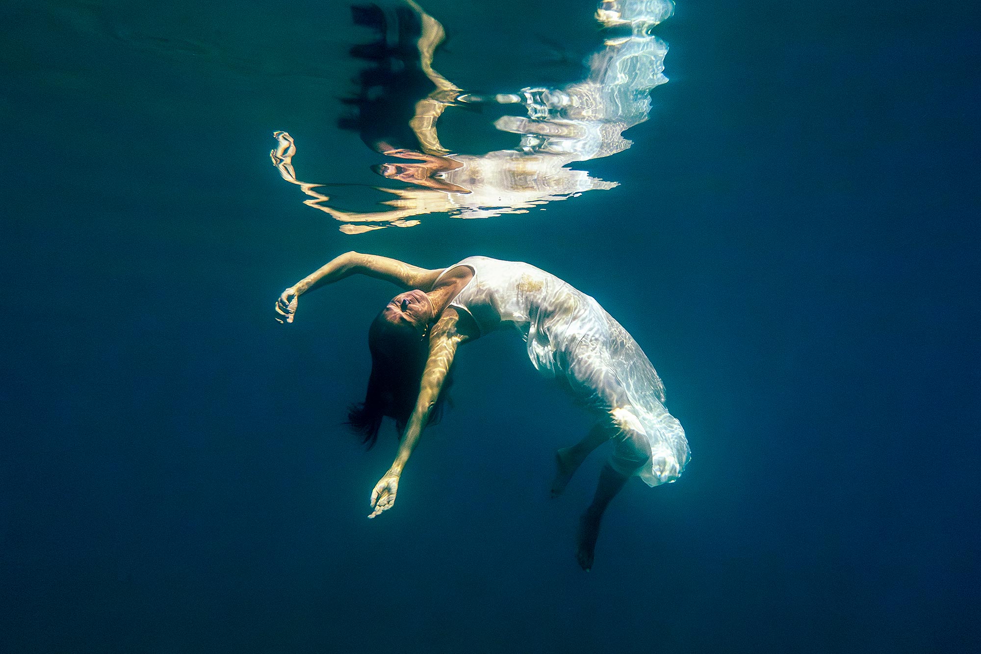 Underwater fine art photograph of a woman floating freely in deep blue water, wearing a white dress, her body gently arched as her body reflects across the surface above.